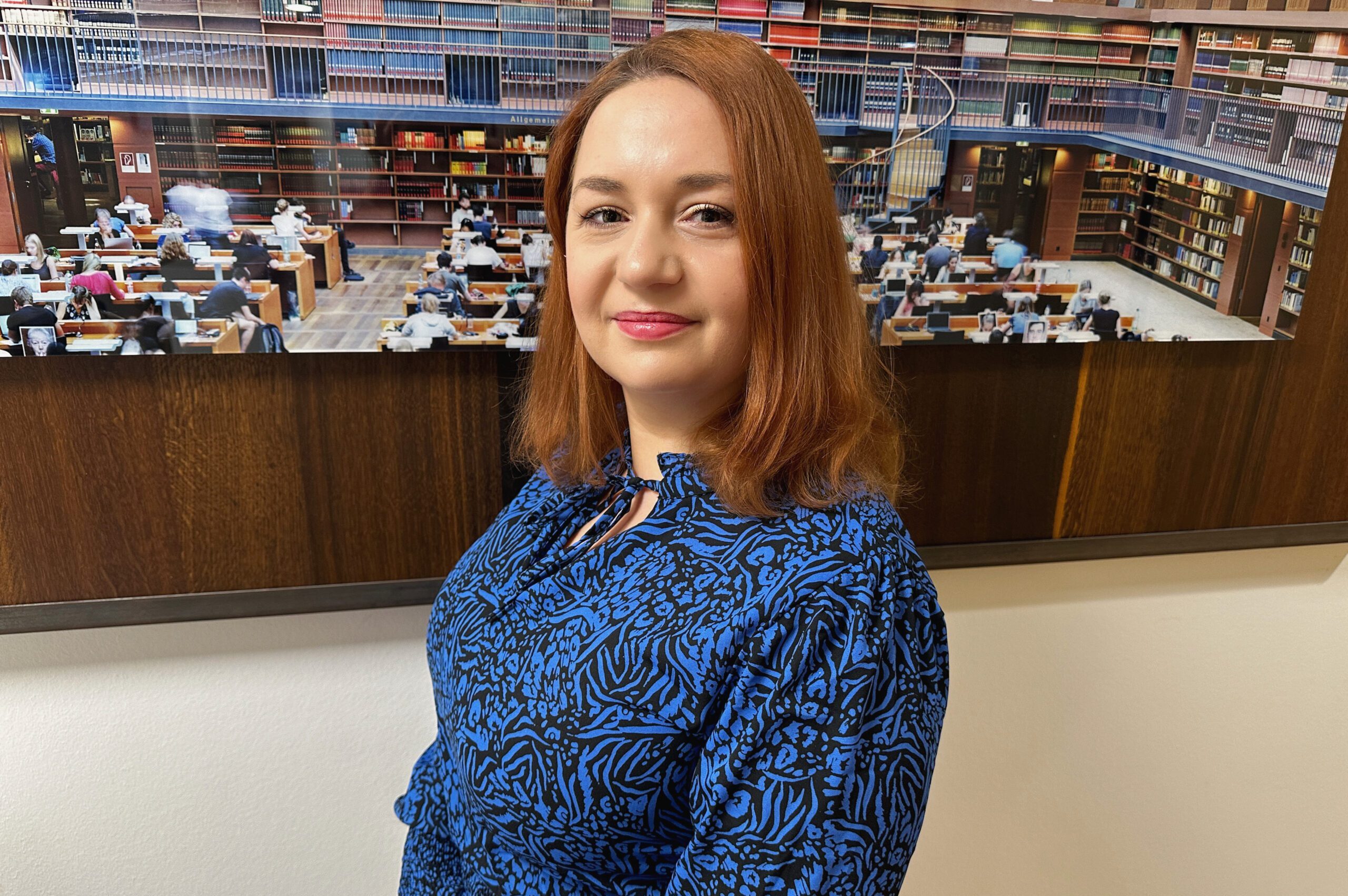 Rechtsanwalts­- & Notarfachangestellte Justizfachangestellte Jessica Plomitzer vor einer Fotowand mit Bibliotheksszene.